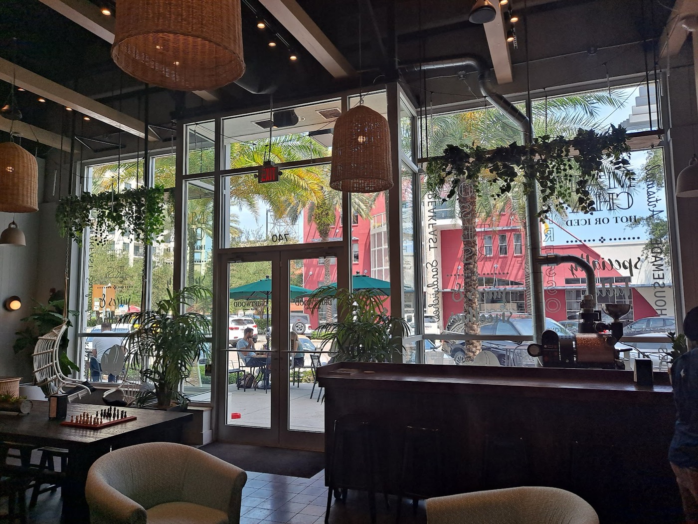 A plant-filled lounge with woven pendants and a hanging rattan chair, looking out tall windows onto Central Avenue.