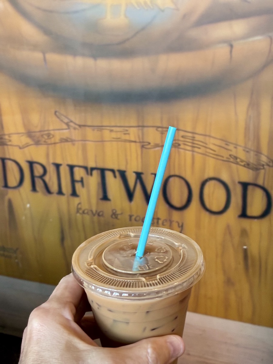 An iced latte held in front of Driftwood’s painted driftwood mural.